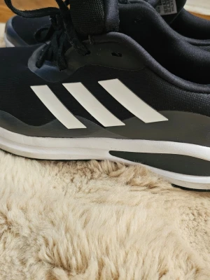Svarta Adidas sneakers med vita detaljer - Snygga svarta sneakers från Adidas med klassiska vita ränder på sidan och vit sula. Skorna har en låg profil och är tillverkade i textil och syntetmaterial. Perfekta för dig som gillar sportig stil och vill ha något som sticker ut.