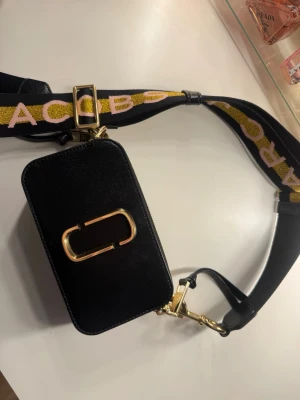 Svart axelväska Marc Jacobs - Snygg svart axelväska från Marc Jacobs med bred axelrem i svart, gult och rosa med logga. Väskan har guldiga metalldetaljer, två dragkedjor och en vit mittpanel. Perfekt för att lyfta din outfit.