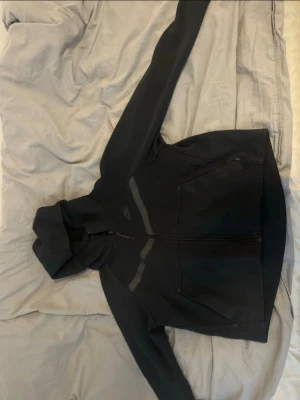Svart hoodie från Nike med dragkedja - Svart hoodie från Nike med hel dragkedja framtill och två stora fickor. Snygga detaljer med diskreta ränder på bröstet. Klassisk huva och sportig look, perfekt för chill eller träning. Tillverkad i mjukt material för extra komfort. Den är dock lite kort i storlek, men använd 2 gånger endast.