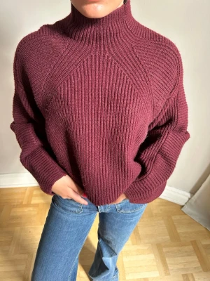 Vinröd stickad polotröja - Mysig vinröd stickad tröja med hög krage och snyggt ribbstickat mönster. Tröjan har en avslappnad passform och långa ärmar, perfekt att matcha med jeans för en chill look.