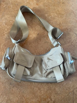 Beige axelremsväska med fickor från divided H&M - Snygg beige handväska i nylon med justerbar axelrem. Väskan har två framfickor med lock och spännen samt dragkedjeförsedda sidofickor. Perfekt för dig som gillar praktiska detaljer och en sportig look.