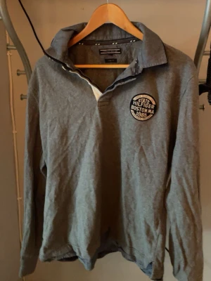 Grå rugbytröja från Tommy Hilfiger - Säljer en grå långärmad rugbytröja från Tommy Hilfiger med krage och knappslå. På bröstet finns ett runt märke med texten 'State Hilfiger Boston MA 1985'
