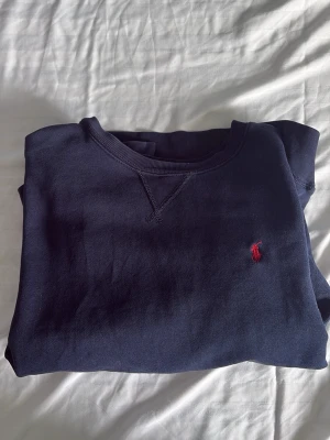 Mörkblå crewneck från Ralph Lauren - Mörkblå sweatshirt från Ralph Lauren med klassisk rund halsringning och röd broderad logga på bröstet. Tröjan har ribbade muddar vid ärmslut och nederkant, perfekt för en avslappnad och stilren look. Litet hål vid handleden högern. Storlek 14-16 år 