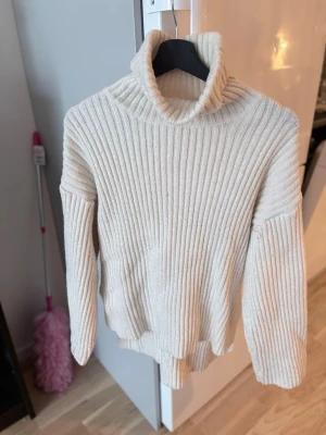 Beige ribbstickad polotröja - Säljer en beige ribbstickad polotröja med hög krage och långa ärmar. Tröjan har en avslappnad passform och slitsar i sidorna vid nederkanten. Perfekt för lager-på-lager och enkel att matcha med jeans eller kjol.