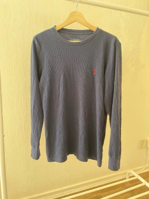 Blå långärmad tröja från Polo Ralph Lauren - Mörkblå långärmad tröja från Polo Ralph Lauren med diskret ribbad struktur och röd broderad logga på bröstet. Tröjan har rund halsringning och är tillverkad i ett mjukt, bekvämt material.