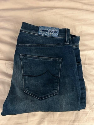 Mörkblå jeans från Jacob Cohën - Snygga mörkblå jeans från Jacob Cohën med klassisk femficksdesign och diskreta slitningar. Jeansen har en stilren passform och detaljer som logoknapp och patch baktill. Perfekta för dig som gillar exklusiva och handgjorda jeans i högkvalitativ denim.
