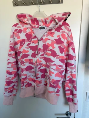 BAPE rosa camo zip hoodie - Säljer en ikonisk BAPE zip hoodie med rosa camo-mönster och hel dragkedja. Hoodien har broderade detaljer och apansikte på huvan samt färgglada bokstäver bak. Mjuka muddar och fickor framtill. Perfekt statement-plagg för streetwear-stilen.