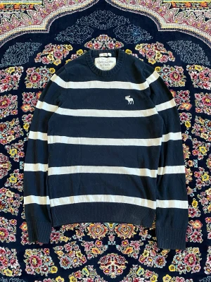 Randig stickad tröja Abercrombie & Fitch - Mörkblå stickad tröja med breda vita ränder från Abercrombie & Fitch. Tröjan har rund halsringning, långa ärmar och en broderad älg-logga på bröstet. Ribbstickade muddar vid ärmslut och nederkant. Perfekt för en avslappnad stil.
