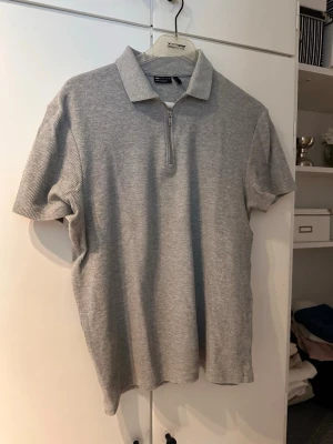 Grå ribbad polotopp med dragkedja - Säljer en ljusgrå ribbad polotopp från med korta ärmar och en snygg dragkedja framtill. Toppen har en klassisk krage och är gjord i mjuk bomullsmix som känns skön mot huden. Perfekt till jeans eller kjol för en clean look.