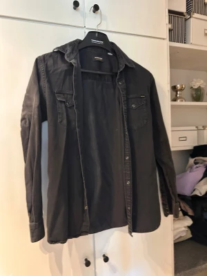 Svart långärmad skjorta från Jack & Jones - Svart skjorta från Jack & Jones i bomull med klassisk krage, bröstfickor med knappar och lång ärm. Skjortan har en avslappnad passform och är perfekt att styla lager på lager.
