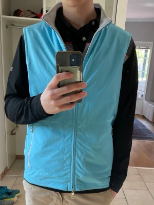 Peter Millar väst - Snygg ljusblå och turkos väst med hel dragkedja framtill och två vita dragkedjefickor. Västen har en hög krage med vit insida och är tillverkad i ett lätt, vindtätt material. Perfekt för golf och annan sport och ger en fräsch look till din outfit.
