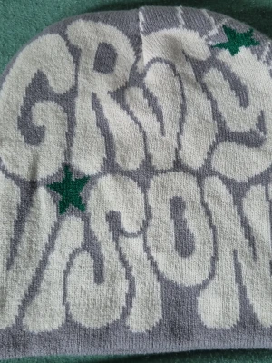 Gråvit mössa med gröna stjärnor - Säljer en stickad mössa i grått och vitt med grafiskt mönster och två gröna stjärnor. Mössan har en rundad form och är tillverkad i mjukt material, perfekt för dig som gillar streetstyle och vill ha en unik accessoar.