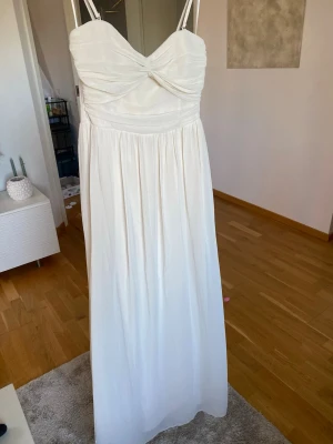 Klänning  - Elegant beige långklänning med tunna axelband och twistad detalj vid bysten. Klänningen har ett draperat liv och ett flytande, plisserat kjolparti. Tillverkad i ett lätt, skirt tyg som ger en romantisk känsla.