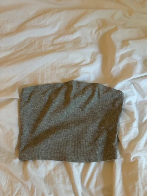 Grå ribbad tubtopp - Säljer en enkel och stilren grå ribbad tubtopp. Perfekt att matcha med jeans eller kjol för en clean look. Toppen är ärmlös och har en bandeau-form som sitter snyggt på plats.