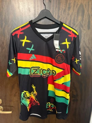 Ajax x Bob Marley Adidas tröja - Säljer en limited edition Ajax x Bob Marley tröja från Adidas. Svart bas med färgglada ränder i rött, gult och grönt, samt grafiska detaljer och Ziggo-logga. V-ringad modell i lätt och ventilerande material, perfekt för fotboll eller streetwear.