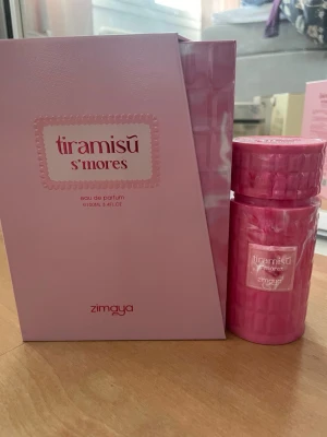 Zimaya Tiramisu S'mores Eau de Parfum 100ml - Tiramisu S'mores Eau de Parfum från Zimaya kommer i en snygg rosa flaska med matchande ask. Doften är söt och gourmand-inspirerad, perfekt för dig som gillar unika och lekfulla parfymer. Flaskan är tillverkad i plast med ett mönstrat lock och etikett framtill. Endast testad!