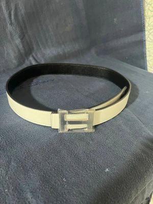 Vitt skärp med H spänne. H’e’r’m’s’e - Snyggt vitt skärp med ett unikt transparent spänne i plast. Bältet har en stilren design och insidan är svart, vilket ger en cool kontrast. Perfekt accessoar för att lyfta din outfit och ge den en modern touch.