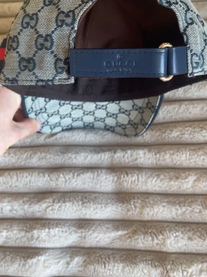 Gucci keps med GG-mönster - Snygg keps från Gucci i beige och marinblått med klassiskt GG-monogram över hela kepsen. Justerbar rem bak i mörkblått läder och röd-blå rand på sidan. Tillverkad i canvasmaterial med detaljer i läder. Perfekt för dig som vill ha en exklusiv och trendig look.