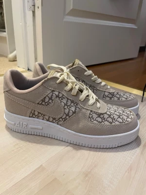 Nike Air Force 1 beige med mönster - Nike Air Force 1 sneakers i beige mocka och textil med unikt D*or-inspirerat mönster på tå och sidor. Skorna har vita sulor, krämfärgade skosnören och klassisk Air Force 1-design. Perfekta för dig som vill sticka ut med en exklusiv look.