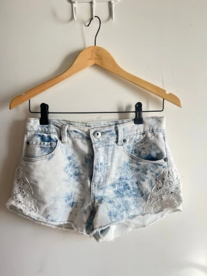 Blåvit jeansshorts med spetsdetaljer - Säljer ett par blåvita jeansshorts med snygg batikmönster och spetsdetaljer på sidorna. Shortsen har råa kanter och fem klassiska fickor. Perfekta för sommaren och ger en avslappnad men trendig look.