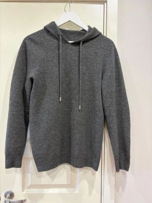 Kashmir hoodie - En stilren grå hoodie med huva och dragsnören. Tillverkad i en mjuk och skön kashmirmix som är väldigt skön och varm. Klassisk passform med ribbade muddar vid ärmslut och nederkant. Perfekt för dig som gillar en clean och enkel look. Köpt på rogé clothing och säljs nu för den inte längre används.