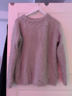 Beige fluffig stickad tröja - Mysig beige stickad tröja med fluffig, lurvig textur. Tröjan har rund halsringning och långa ärmar. Perfekt för dig som gillar en mjuk och trendig look. Materialet känns som en blandning av syntet och ull för extra komfort.