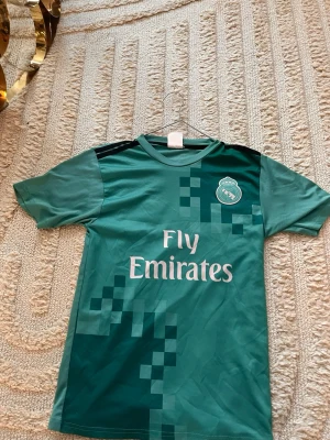 Real Madrid Ronaldo matchtröja grön - Snygg grön Real Madrid matchtröja med Ronaldo 7 på ryggen och Fly Emirates-tryck på framsidan. Tröjan har korta ärmar, svart detalj på axlarna och pixelmönster i olika gröna nyanser. Perfekt för fotbollsfans som vill sticka ut på planen.
