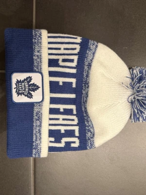 Toronto Maple Leafs blåvit mössa - Säljer en blåvit mössa från Toronto Maple Leafs med uppvikt kant och broderad logga framtill. Mössan har ett stort texttryck runtom och en matchande blåvit tofs på toppen under kalla dagar.