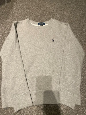 Grå sweatshirt från Polo Ralph Lauren - Klassisk grå sweatshirt från Polo Ralph Lauren med rund halsringning och den ikoniska blå broderade loggan på bröstet. Storlek S
