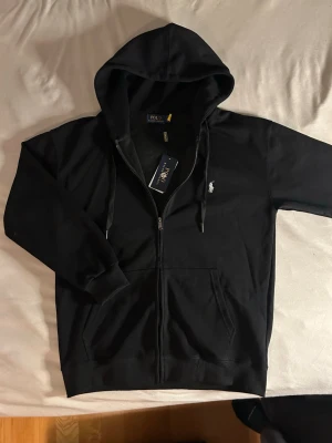 Svart hoodie från Polo Ralph Lauren - Snygg svart hoodie från Polo Ralph Lauren med dragkedja framtill och klassisk vit logga broderad på bröstet. Tillverkad i mjukt bomullsmaterial som känns skönt mot huden. Står L men passar s/m då den är kortare i armarna, aldrig använt den