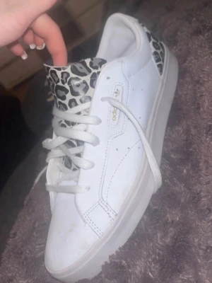 Vita Adidas sneakers med leoparddetaljer - Säljer ett par vita Adidas sneakers med snygga leopardmönstrade detaljer på plösen och hälen. Skorna har klassisk låg profil, vita snören och diskret guldfärgad Adidas-logga på sidan. Tillverkade i slätt läder för en clean och trendig look.