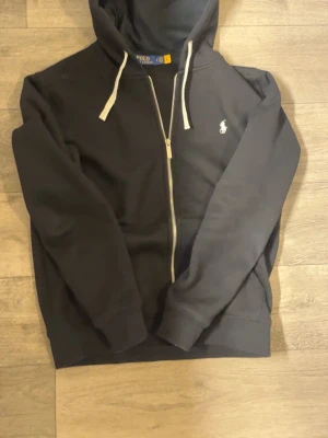  Ralph Lauren zip  hoodie - Snygg svart hoodie från Polo Ralph Lauren med dragkedja framtill och vit broderad logga på bröstet. Hoodien har vita dragsnören i huvan och ribbade muddar vid ärmar och nederkant. Perfekt för en avslappnad och stilren look.