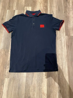 SlimFit Marinblå pikétröja från Hugo - Snygg marinblå pikétröja från Hugo med röda detaljer på krage och ärmslut samt röd logotyp på bröstet. Klassisk pikékrage med knappar framtill. Perfekt för en stilren och avslappnad look.