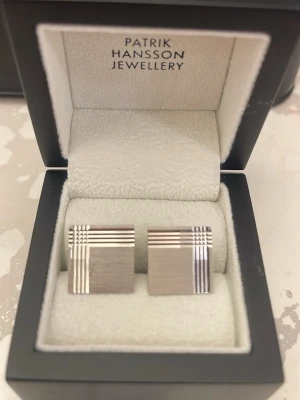 Silvriga manschettknappar Patrik Hansson - Stilrena silvriga manschettknappar från Patrik Hansson Jewellery. Fyrkantig form med borstad yta och dekorativa linjer i blank finish som skapar ett rutmönster i hörnet. Levereras i original ask. Perfekt accessoar för att lyfta skjortan.