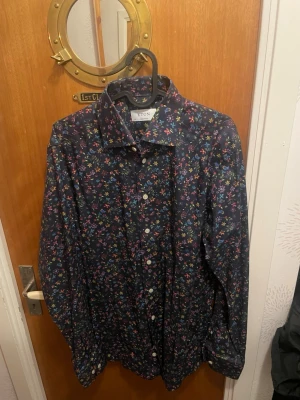 Blommig marinblå skjorta från Eton - Snygg marinblå skjorta från Eton med färgglatt blommigt mönster. Skjortan har klassisk krage, vita knappar och lång ärm. Perfekt för dig som vill sticka ut med stilrena detaljer och skön passform. Materialet är mjuk bomull som känns bekvämt hela dagen. Har använts en eller två gånger och är i storlek 40. Nypris 1200kr +.