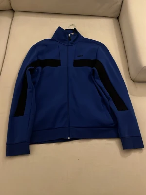 Blå ziptröja från BOSS - Snygg blå ziptröja från BOSS med svarta detaljer längs ärmar och bröst. Tröjan har hög krage, hel dragkedja framtill och en sportig look. Perfekt för dig som gillar stilrena och moderna plagg.