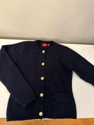 Mörkblå kofta med guldknappar - Stilren mörkblå kofta från ryvls med rund halsringning och sex dekorativa guldknappar framtill. Koftan har långa ärmar, ribbade muddar och en praktisk ficka på ena sidan. Perfekt för dig som gillar klassisk men ändå trendig stil. I mörkblått