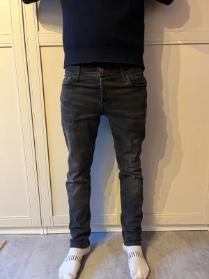 Svarta slim fit jeans herr - Säljer ett par svarta slim fit jeans med klassisk femficksmodell och knappgylf. Jeansen har en något tvättad look och sitter tight över benen för en modern stil. Perfekta för dig som gillar en enkel men snygg look. Jack and Jones jeans
