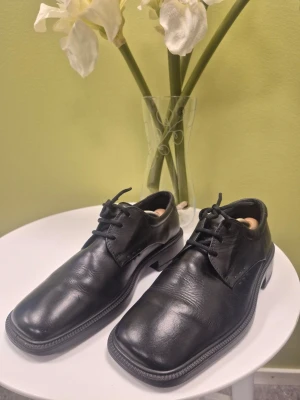 Ecco City svarta loafers i skinn med fyrkantig tå 40 - Stilrena svarta loafers i äkta skinn med fyrkantig tå och klassisk snörning. Skorna har en låg klack och diskreta sömmar längs kanterna för en clean look. Perfekta för dig som gillar minimalistisk och tidlös stil. Stora i storlek,  passar 41, innemått 26cm