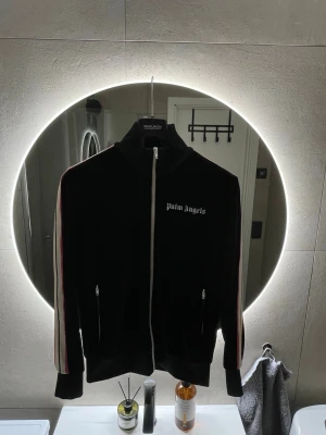 Palm Angels svart ziptröja - Säljer en svart ziptröja från Palm Angels med vita, rosa och orangea ränder längs ärmarna. Tröjan har hög krage, dragkedja framtill och två fickor. Ikonisk Palm Angels-logga på bröstet. Perfekt för trendig look. Tröjan är i bra skick inte använd mycket. Nypris 4795:- köpt på farfetch Storlek L passar M