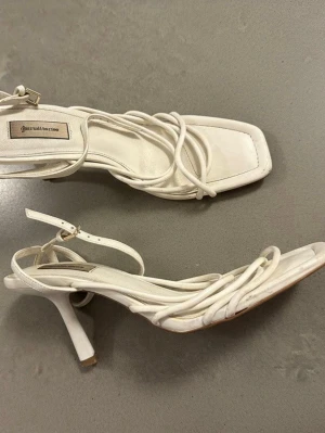 Vita strappy sandaler från Stradivarius - Säljer ett par vita sandaler från Stradivarius med tunna remmar och fyrkantig tå. Sandalerna har en smal klack och justerbart spänne vid ankeln. Tillverkade i konstläder med trendig design som passar perfekt till sommarens outfits. Behövs bara torkas av. 