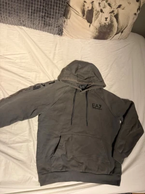 EA7 hoodie - Snygg grå hoodie från EA7 Emporio Armani med svart logga på bröstet och stort tryck längs ena ärmen. Klassisk känguruficka framtill och justerbar huva med snören. Perfekt för en avslappnad och trendig look. Inga håller eller liknade finns på tröjan. 