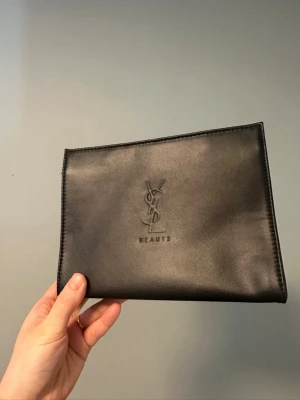 Ny Svart kuvertväska från YSL Beauté sminkväska  - Ny oanvänd. 20*15*7cm. Snygg svart kuvertväska från YSL Beauté i slätt konstläder med präglad logga på framsidan. Enkel och stilren design med rektangulär form, perfekt för att bära det viktigaste. Passar dig som gillar klassiska accessoarer med lyxig känsla.