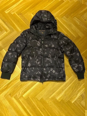 Woolrich jacka - Säljer en pufferjacka från Woolrich med grått camomönster. Jackan har huva, dragkedja framtill och två sidofickor. Muddar vid ärmslut och quiltad design för extra värme. Perfekt för kalla vinterdagar och riktigt snygg streetstil.
