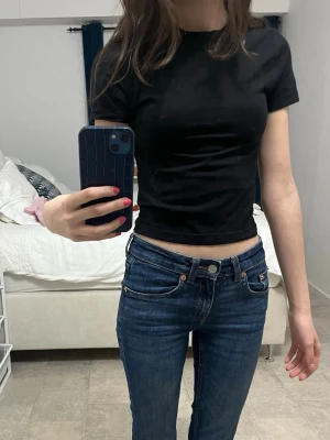 Svart croppad t-shirt - En enkel och stilren svart croppad t-shirt med rund hals och korta ärmar. Perfekt till jeans eller kjol för en clean look. T-shirten är i mjukt bomullsmaterial och har en tight passform som framhäver figuren.