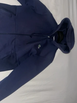 Mörkblå hoodie från Lacoste Sport - Snygg mörkblå hoodie från Lacoste Sport med dragkedja framtill och klassiska krokodilloggan på bröstet. Tillverkad i mjukt bomullsmaterial och har justerbar huva med snören samt fickor på sidorna. Perfekt för en sportig och avslappnad stil.