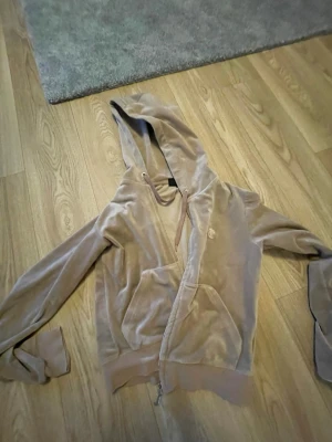 Beige velour hoodie från Juicy Couture - Säljer en beige velour hoodie från Juicy Couture med dragkedja framtill och två fickor. Hoodien har en broderad JC-logga på bröstet och justerbar huva med snören. Perfekt för en chill och trendig look.