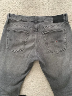 Grå jeans från Tommy Hilfiger - Snygga grå jeans från Tommy Hilfiger med raka ben och klassiska bakfickor. Jeansen har en diskret logga på ena bakfickan och är tillverkade i mjukt denimtyg. Passformen är Slim. Nypris 1400kr.