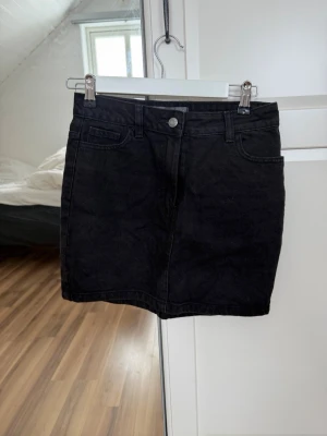 Svart jeanskjol med fickor - Snygg svart jeanskjol med klassisk femficksdesign och bälteshällor. Kjolen har en rak passform och stängs med knapp och dragkedja framtill. Perfekt basplagg att matcha med olika toppar.