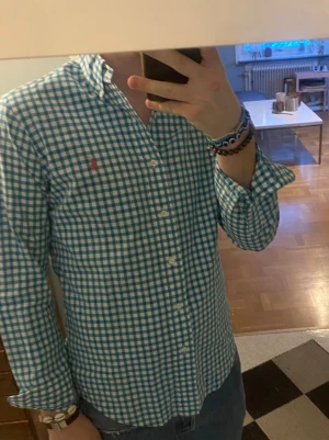 Blårutig skjorta från Ralph Lauren - Säljer en klassisk blårutig skjorta från Ralph Lauren med vit bas och blå rutor. Skjortan har lång ärm, knappar hela vägen och den ikoniska rosa loggan broderad på bröstet. Perfekt till jeans eller chinos för en clean look. Storlek 38 passar S